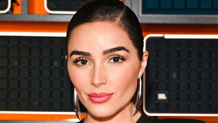Olivia Culpo smiles for the camera sporting a pink glossy lip, slicked-back hairdo and oversized silver earrings.